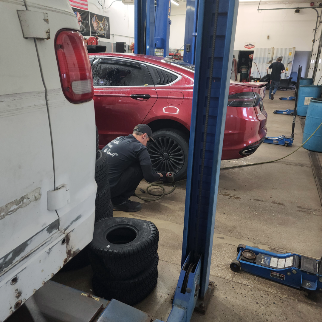Mechanic working on the rear wheel of a red sedan inside an automotive repair shop, with tires, a white van, and blue hydraulic lifts visible in the background.